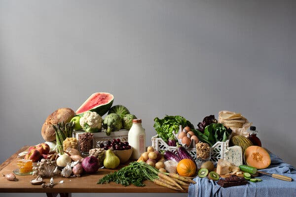 An assortment of food on a wooden table. On the left are some high-FODMAP foods including watermelon, asparagus, onions, honey and a bottle of milk. On the right are some low-FODMAP foods including carrots, eggs, cucumber, lettuce and cantaloupe. Part of the table is covered in a blue tablecloth.