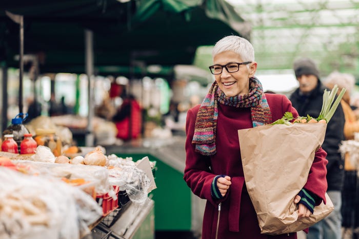 A smiling older woman in a colorful jacket is shopping for healthy groceries at an outdoor farmers market.