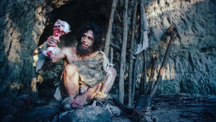 A stock image shows a person wearing animal skins and holding up a meat-covered bone in front of a cave.