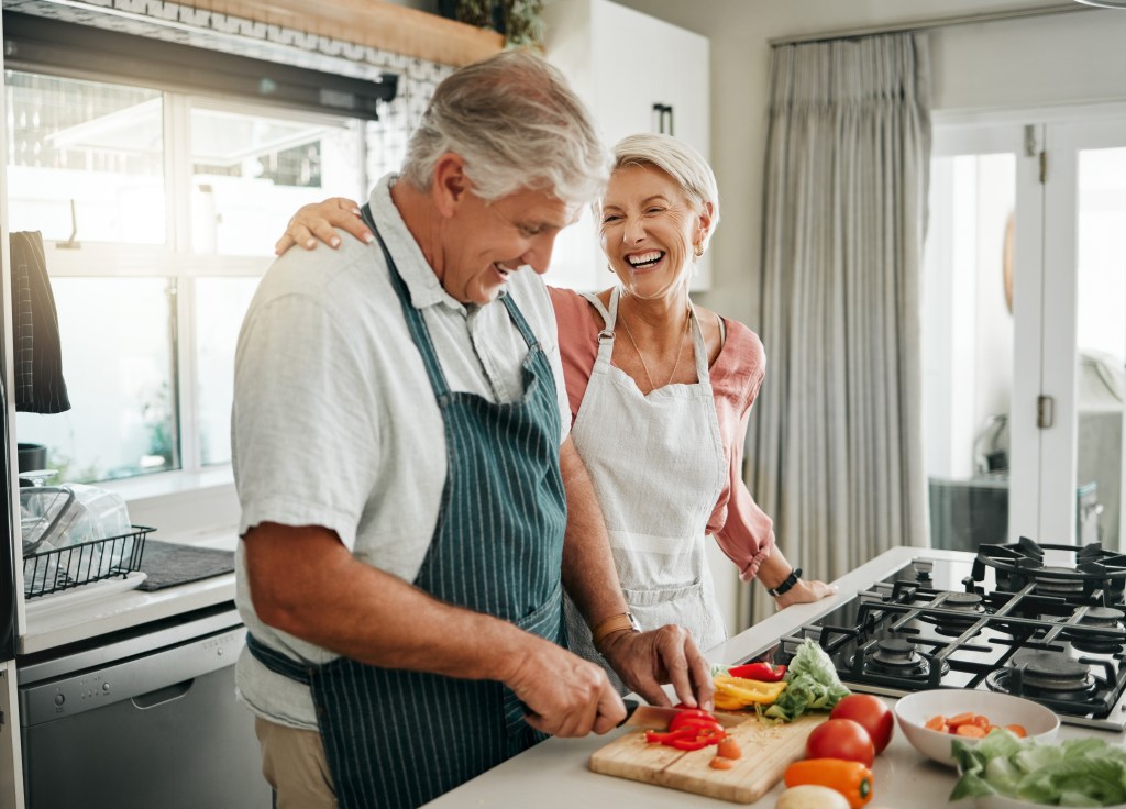 Senior couple, cooking and having fun while preparing a healthy food with vegetables for a vegan meal in the kitchen at home while laughing and having fun. Funny old man and woman helping with dinner.