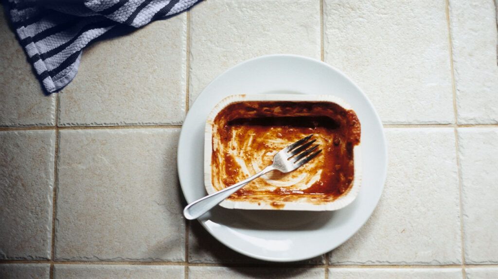 An empty microwave dinner container on a plate with a fork.